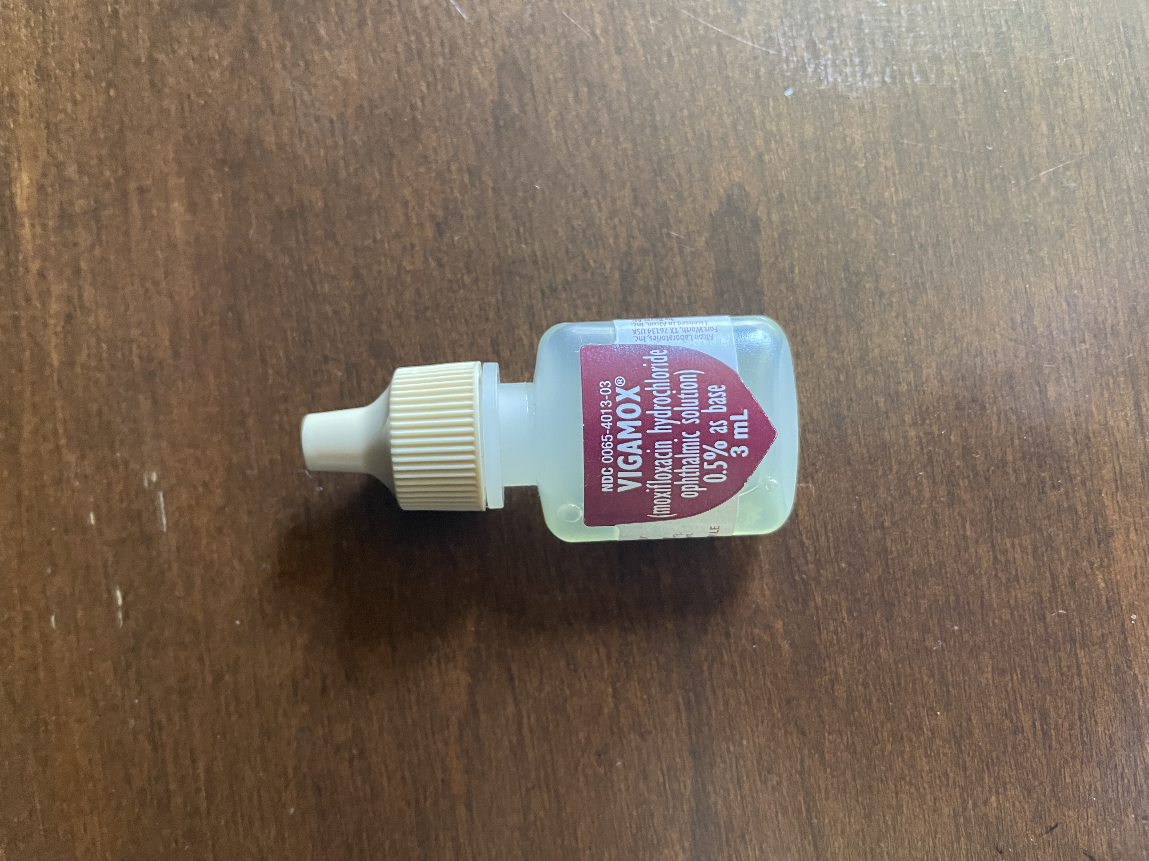 A Vigamox eyedrop bottle on a flat brown surface.